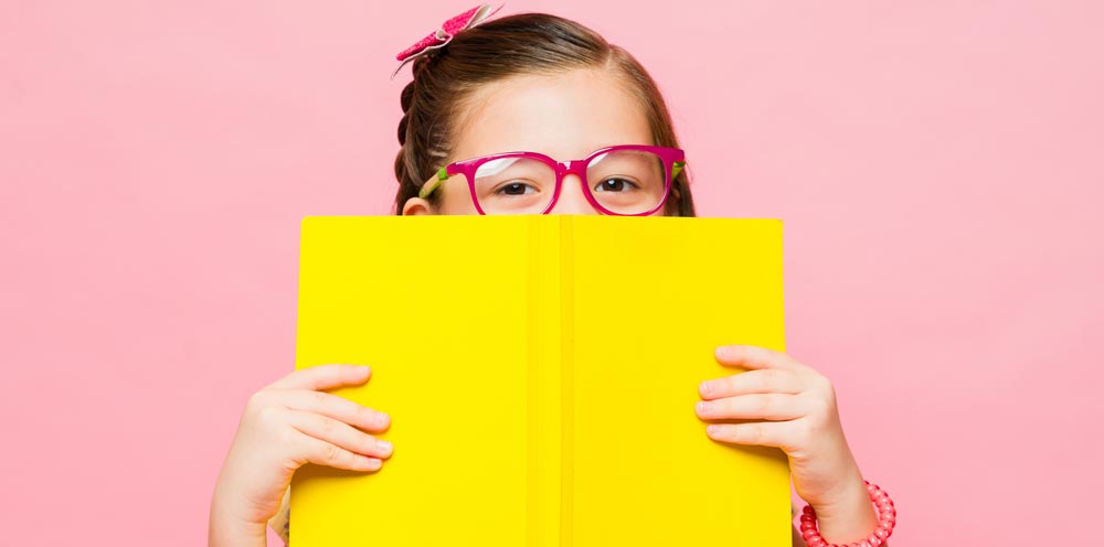 young girl in glasses peaking over a bright yellow book